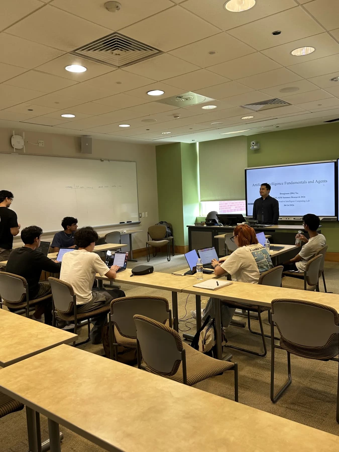 A presenter in front of a slide titled Artificial Intelligence Fundamentals and Agents, with developers working on laptops.