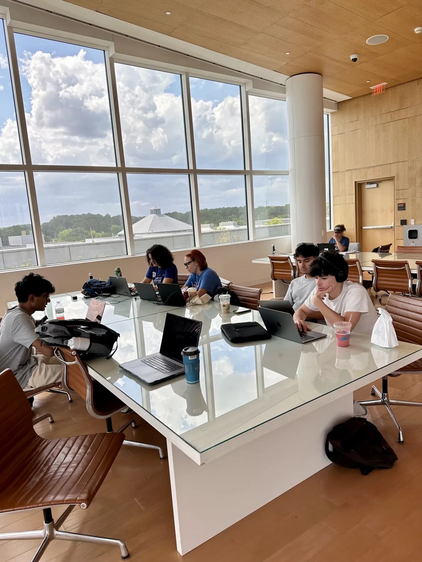 Developers working on laptops around a long glass table beside tall windows.